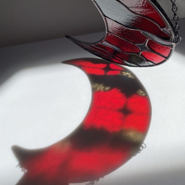 Red and black stained glass cloudy moon suncatcher with red and gray refractions on the ground.